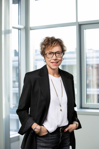 A woman wearing glasses and a black blazer, standing indoors by large windows with hands in pockets and a light-colored shirt.